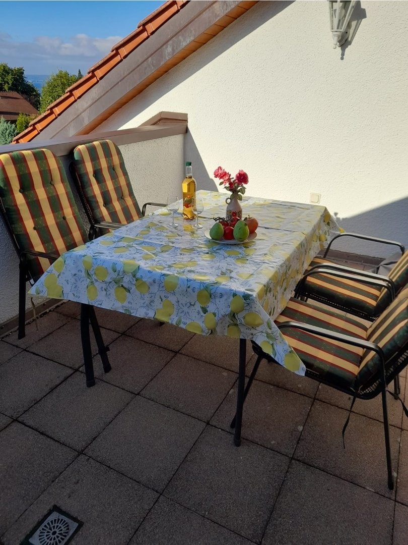Balkon mit Tisch, Stühlen, Blick auf Bodensee und Berge, sonniges Wetter.