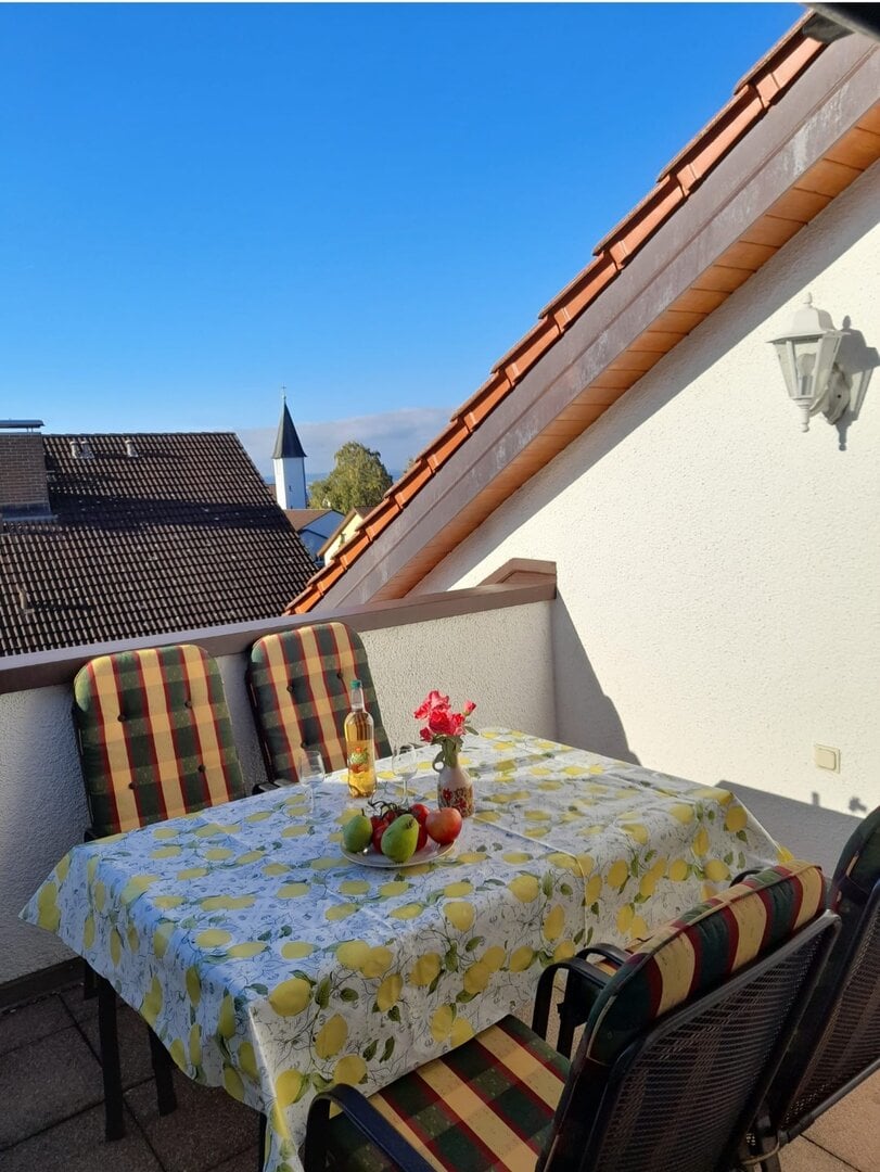 Balkon mit Tisch, Stühlen und Blick auf Bodensee und Berge bei schönem Wetter.
